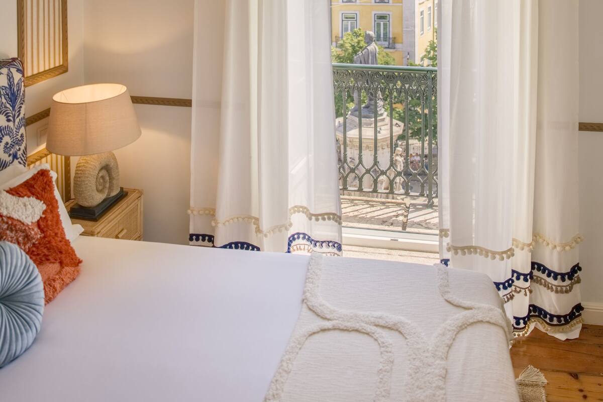 Une chambre à coucher avec un grand lit, un lustre et une vue sur les bâtiments à travers de fines rideaux.