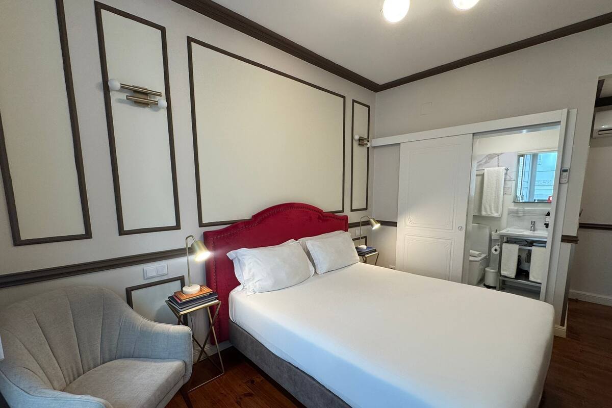 Une chambre d’hôtel avec un grand lit, une table de chevet avec une lampe, une chaise et une petite table avec des livres. On peut apercevoir la salle de bain à travers une porte ouverte.