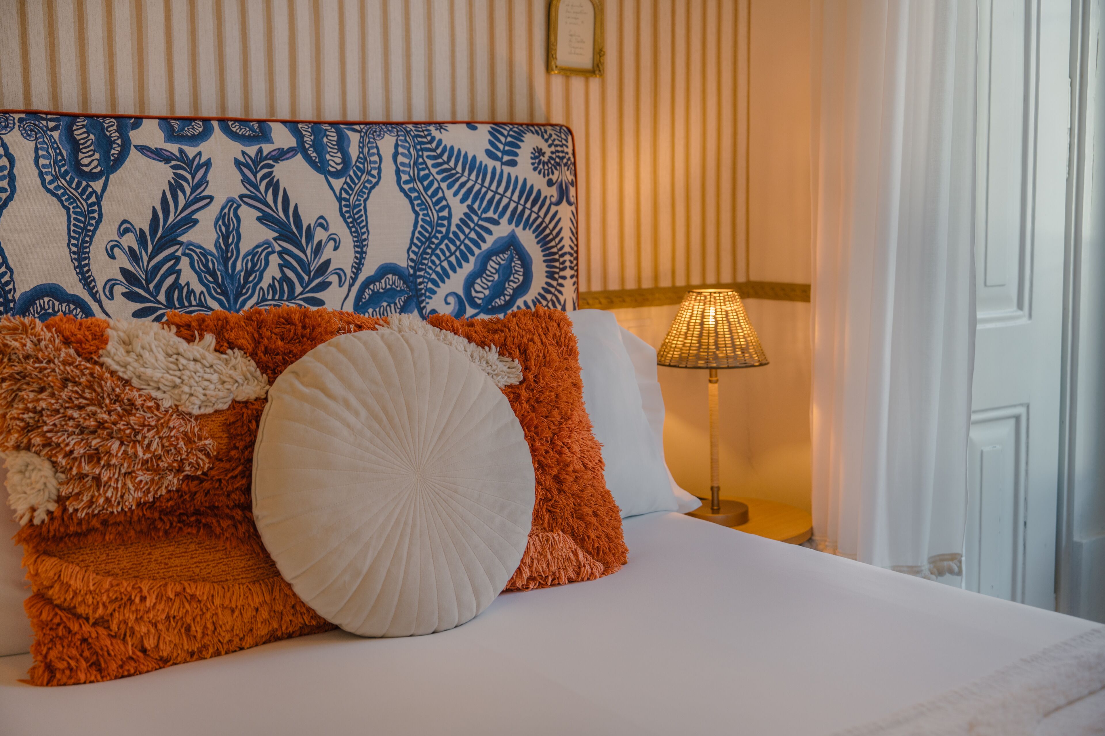 Een net opgemaakt bed met een bekleder met patroon, een lamp en een raam met gordijnen.