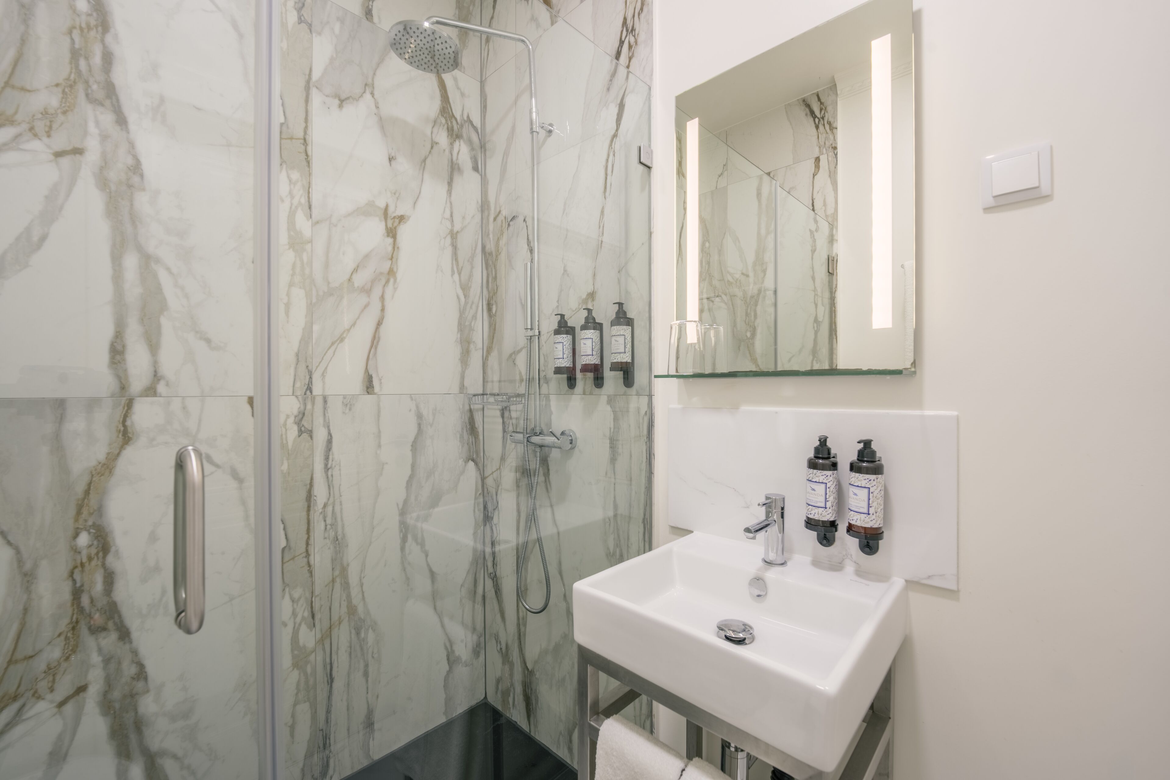 Un baño moderno con ducha de vidrio, lavamanos blanco con dos dispensadores de jabón y un espejo sobre el lavamanos.