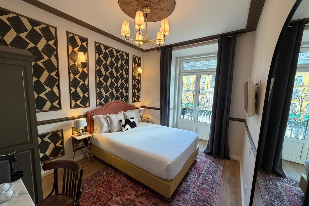 Une chambre d’hôtel avec un lit, un bureau et une chaise, un tapis à motifs et une vue sur les bâtiments par la fenêtre.