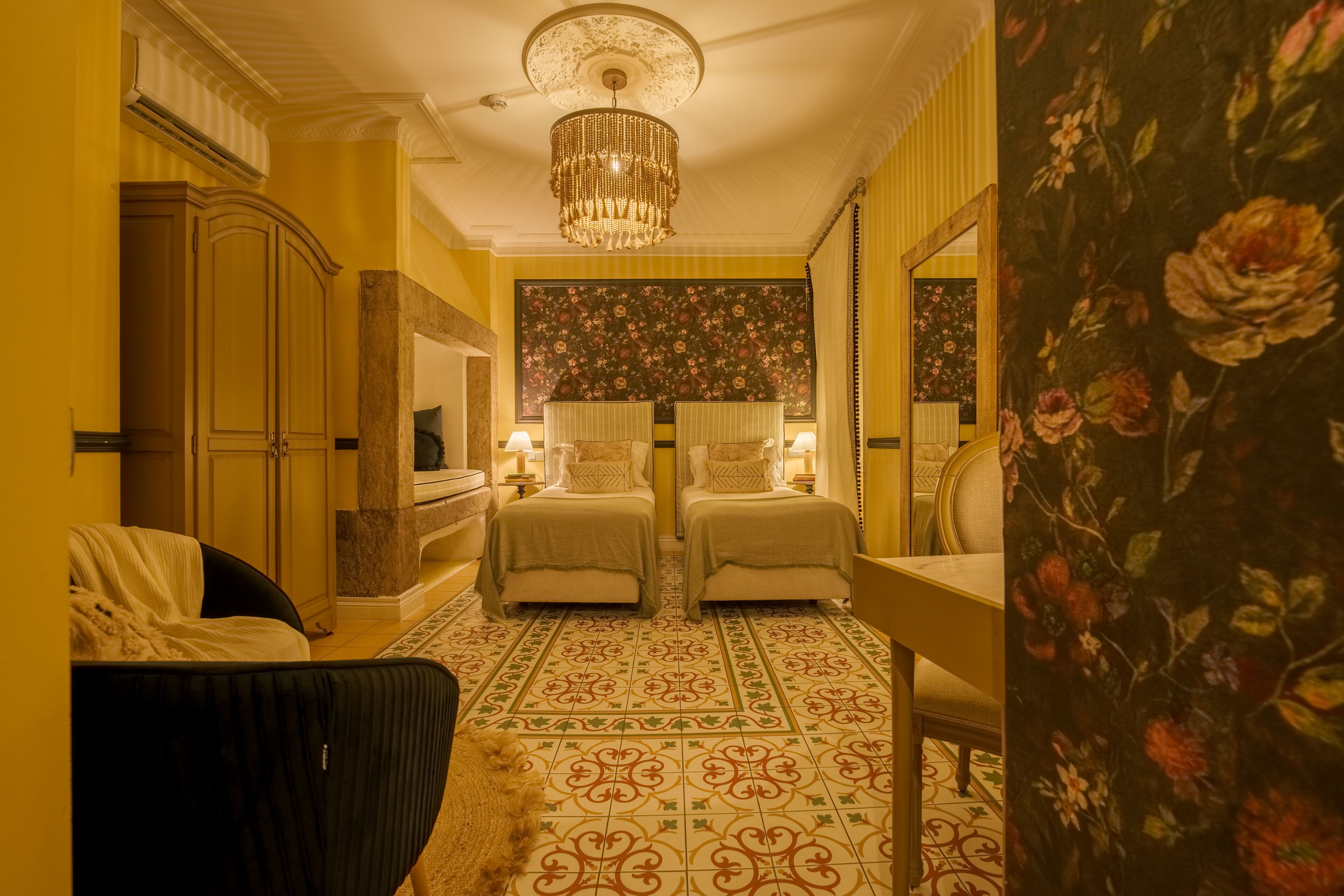A hotel room with two beds, a large mirror, a chandelier, and a floral wallpaper.