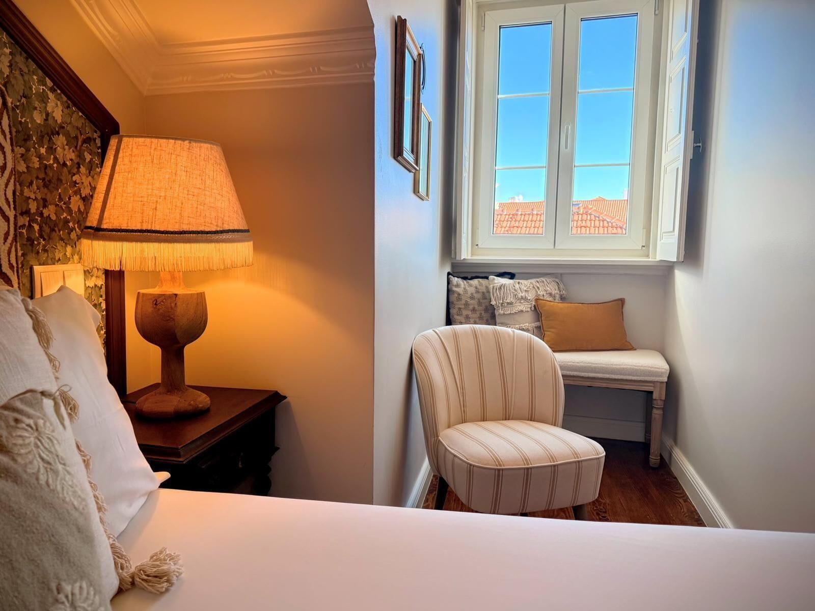 A bedroom with a bed, a wooden nightstand with a lamp, a chair, a window with a view of buildings, and a wall with framed pictures.