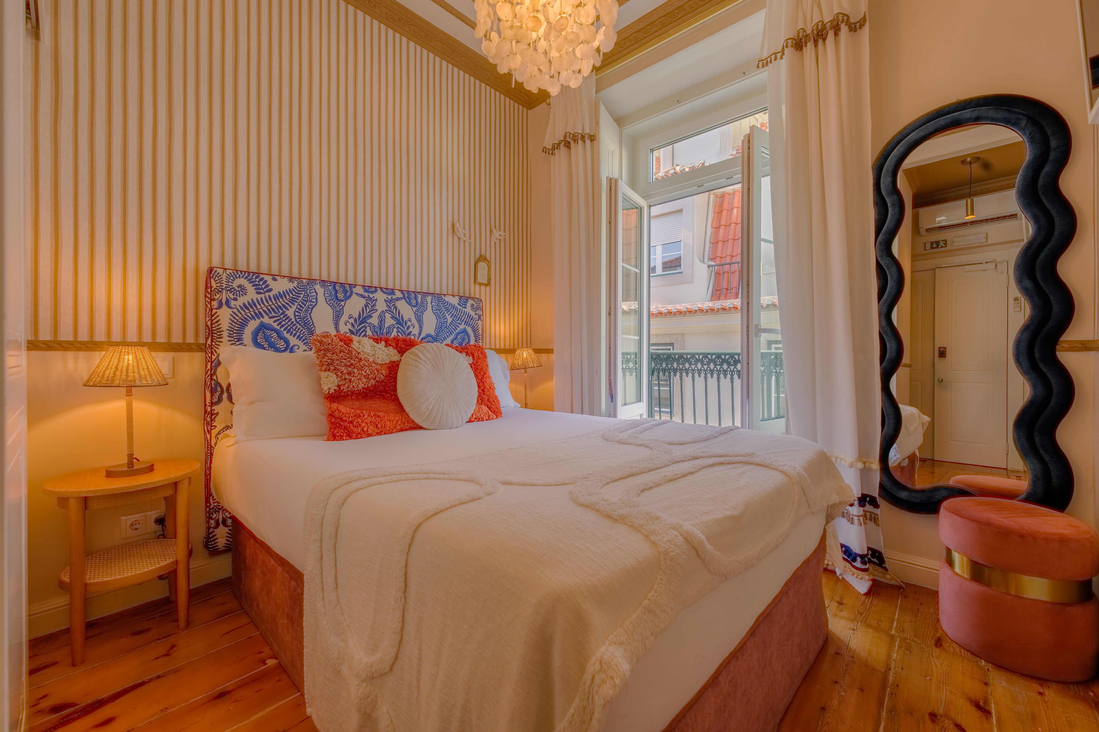 A hotel room with a large bed, a bedside table with a lamp, a mirror, and a view of the outside balcony.