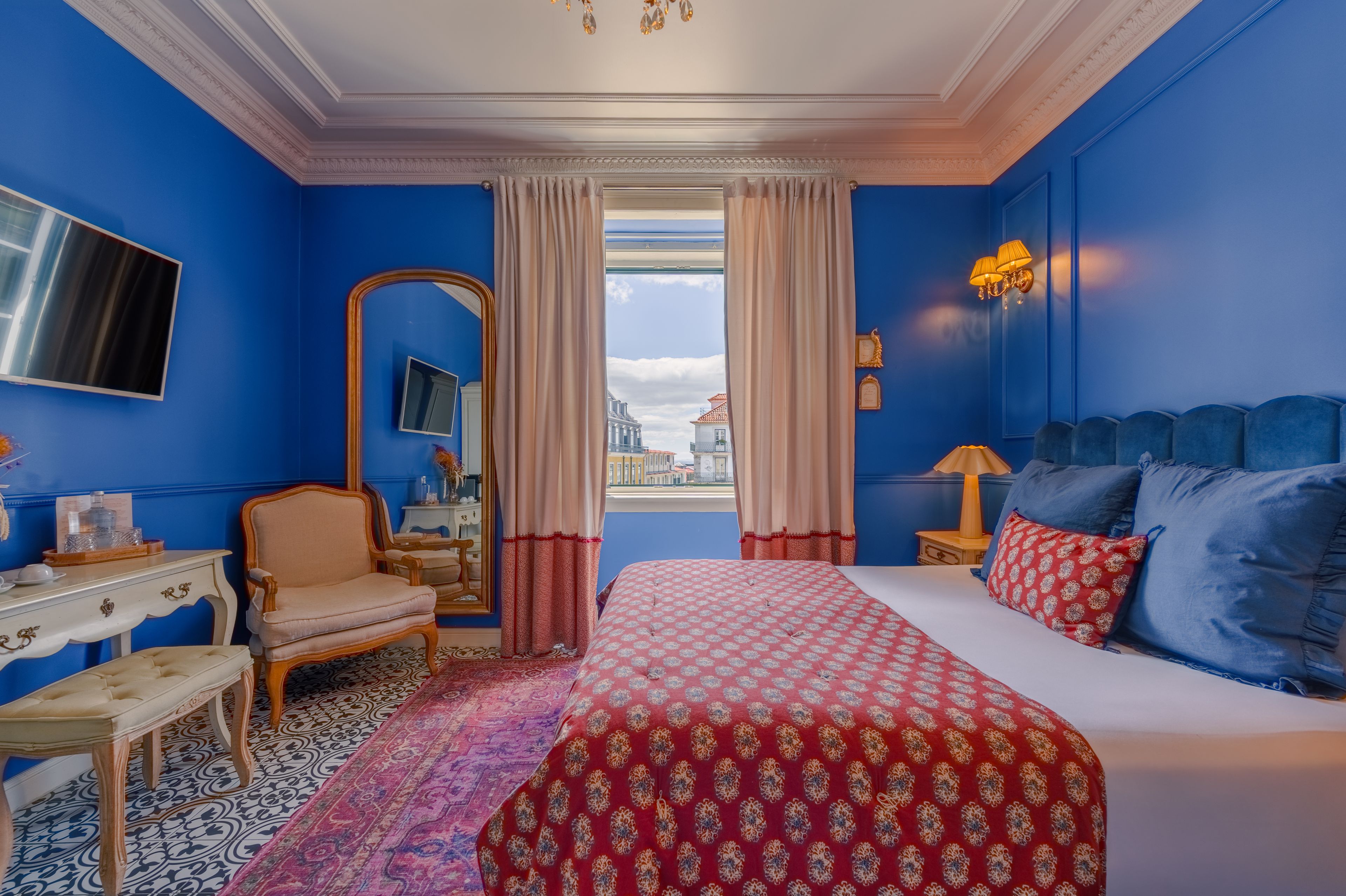 Ein Hotelzimmer mit einem großen Bett, einer Sitzecke mit einem Sessel, einem Schreibtisch, einem Spiegel und Blick auf die Stadt.