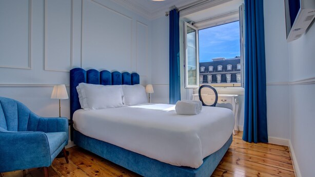 A bedroom with a large bed, a blue armchair, a bedside table with a lamp, and a view of buildings through a window. - Solar dos Poetas (Lisbon)