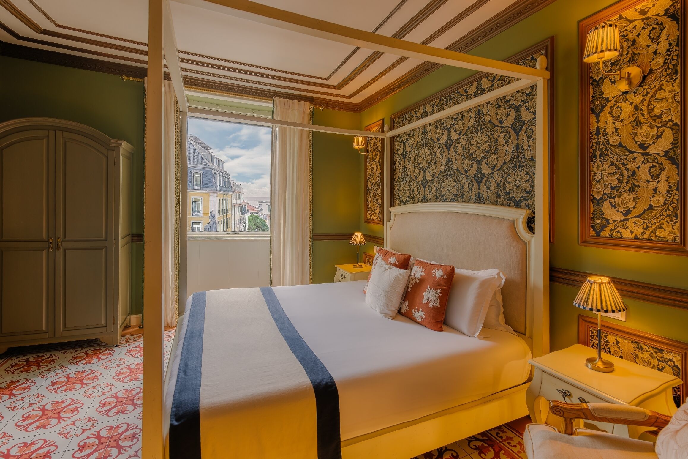 A four-poster bed with a canopy, a nightstand with a lamp, and a view of a cityscape through a window.