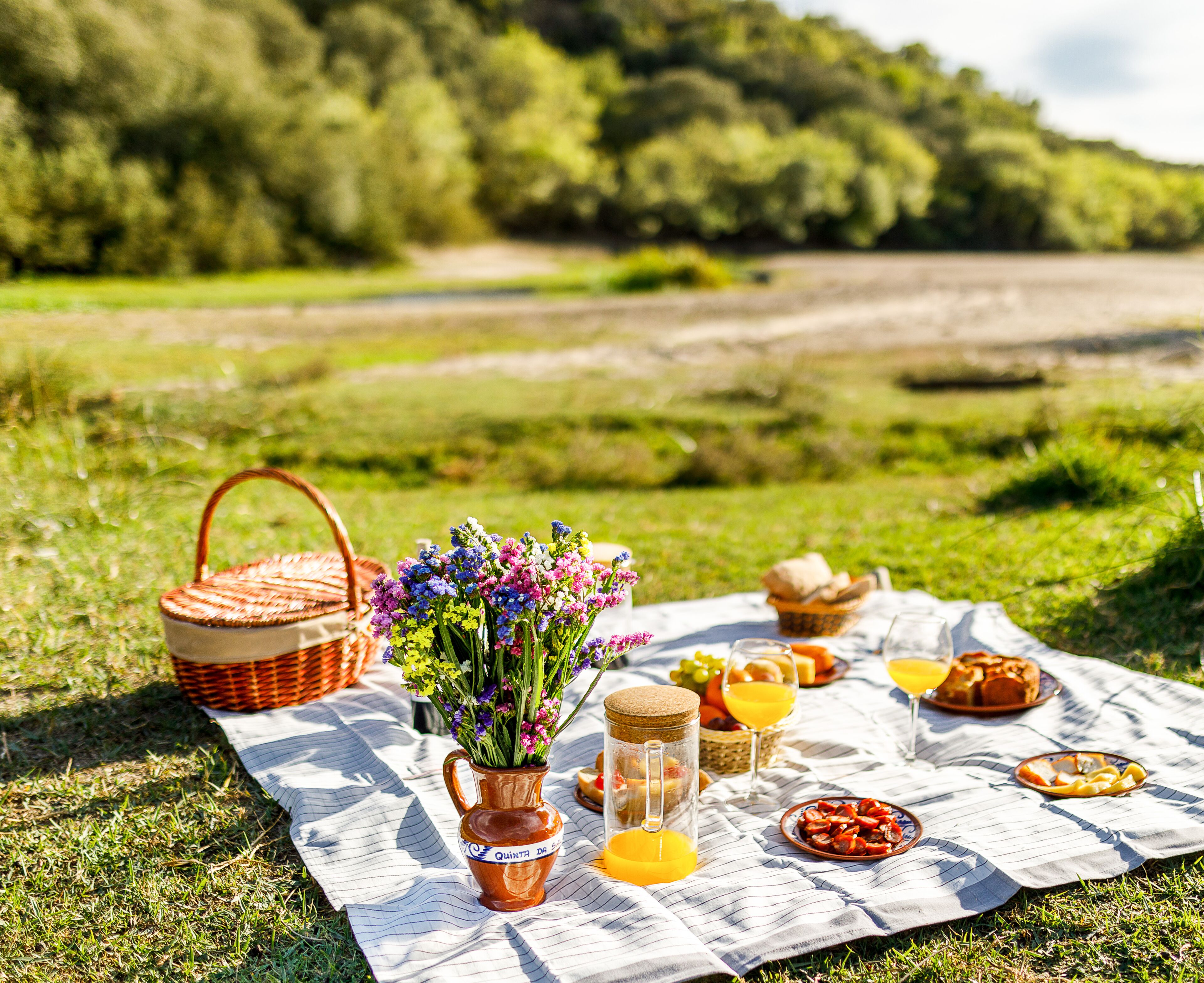 Un montaje para picnic con una cesta, una jarra de flores, un frasco de mermelada y una manta sobre un campo de césped.