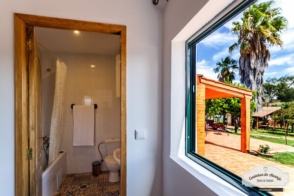Un baño con ducha, inodoro y ventana con vistas a un patio amueblado y palmeras.
