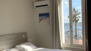 Quarto de hotel com uma cama grande, um ventilador de teto, um ar condicionado, uma janela com vista para o mar e barcos, e um quadro na parede.