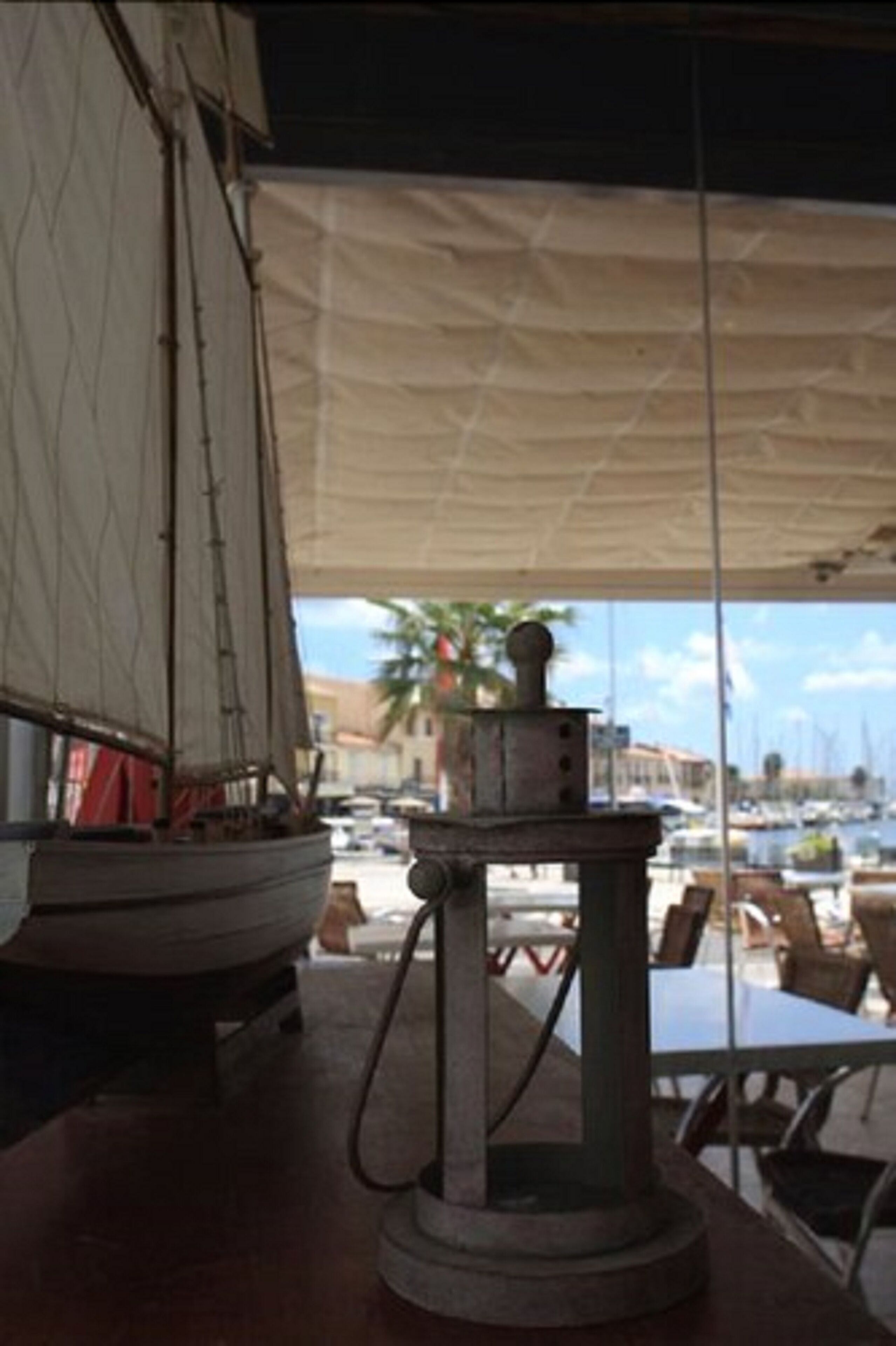Un velero en miniatura sobre una mesa, bajo una toldilla, con vista a un puerto deportivo y embarcaciones al fondo.