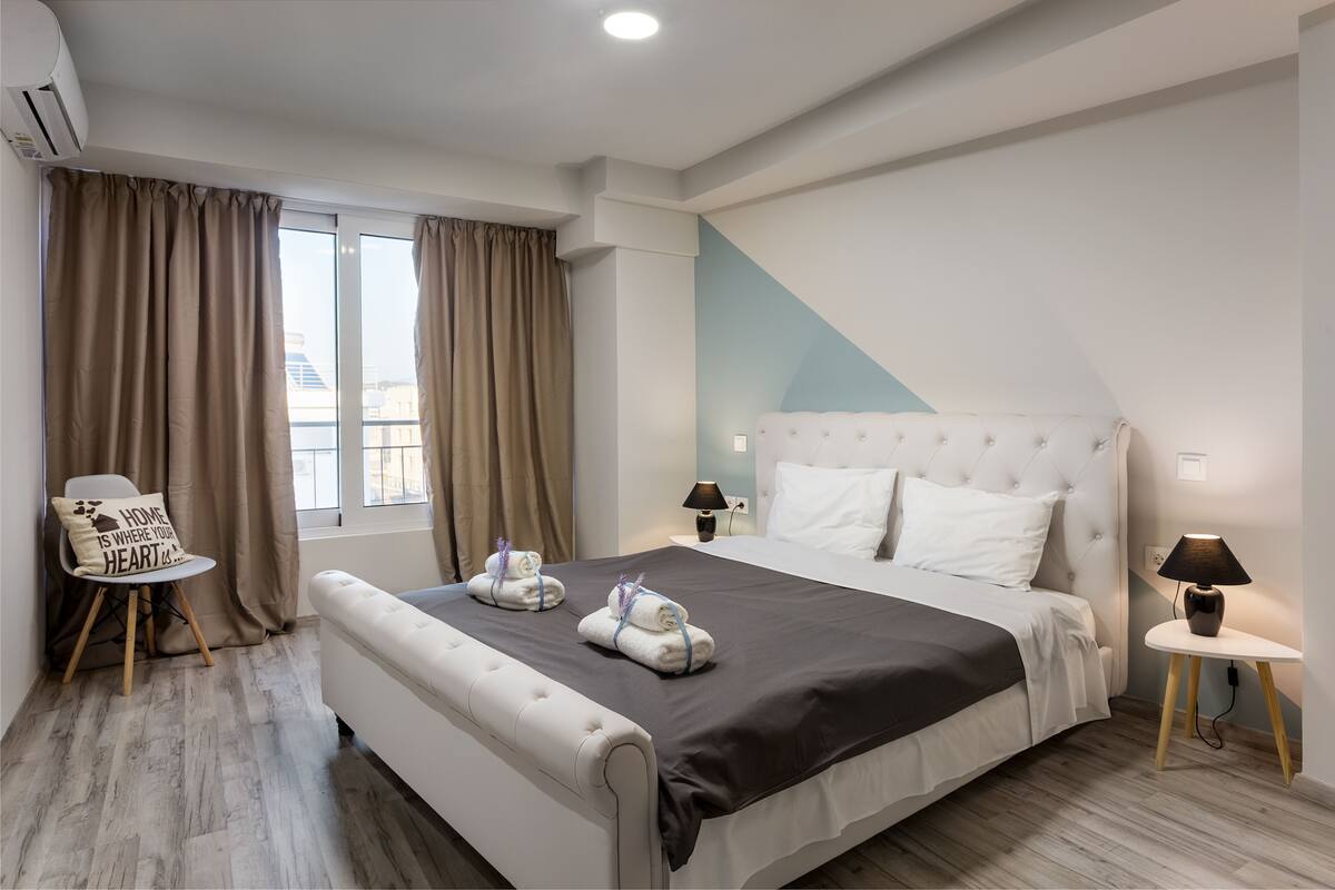 Una habitación de hotel moderna con una cama grande, dos mesitas de noche, una silla y vistas a la ciudad.