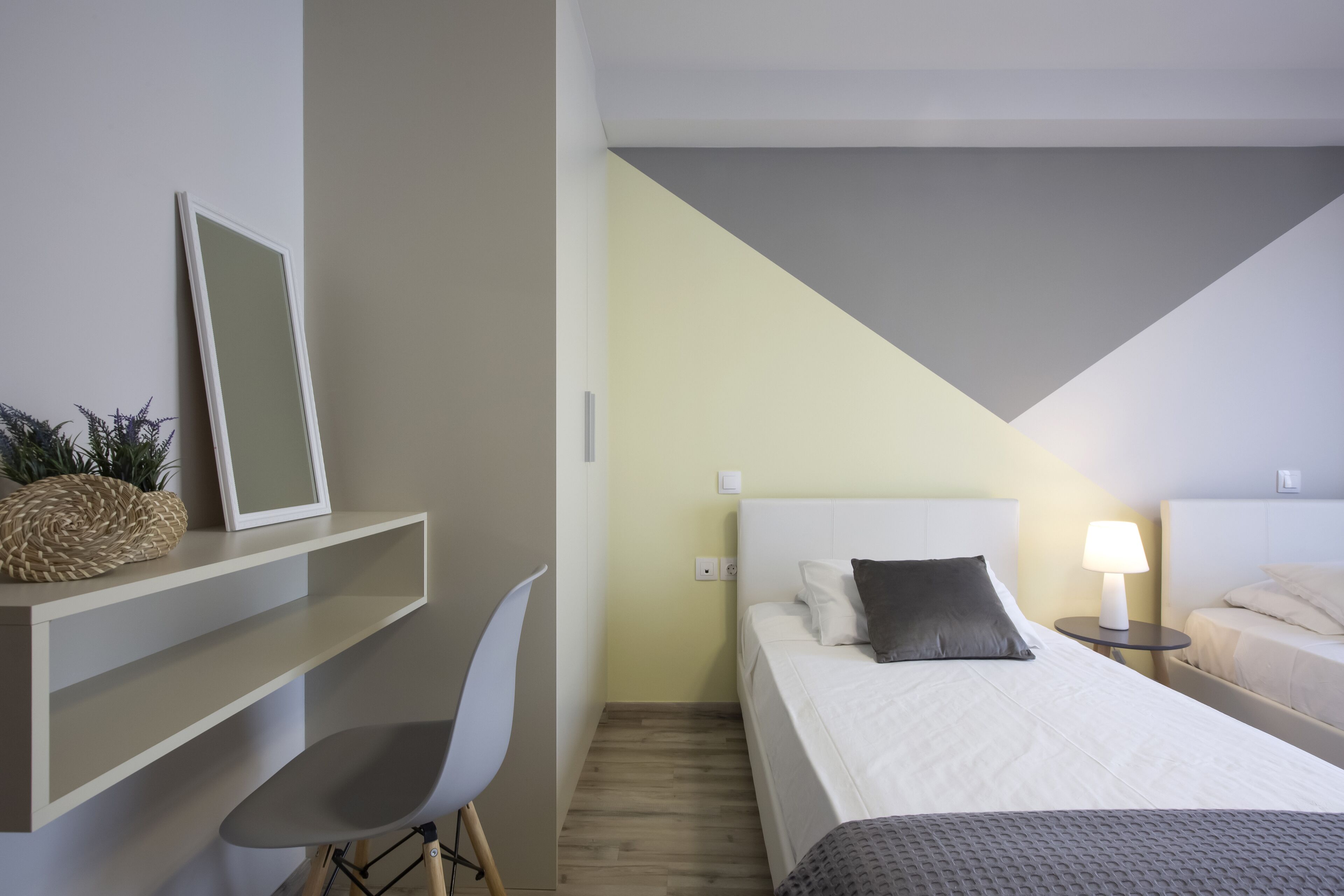 Ein modernes Hotelzimmer mit Bett, Nachttisch, Lampe, Sessel und Schreibtisch mit Spiegel und Dekorationselementen.
