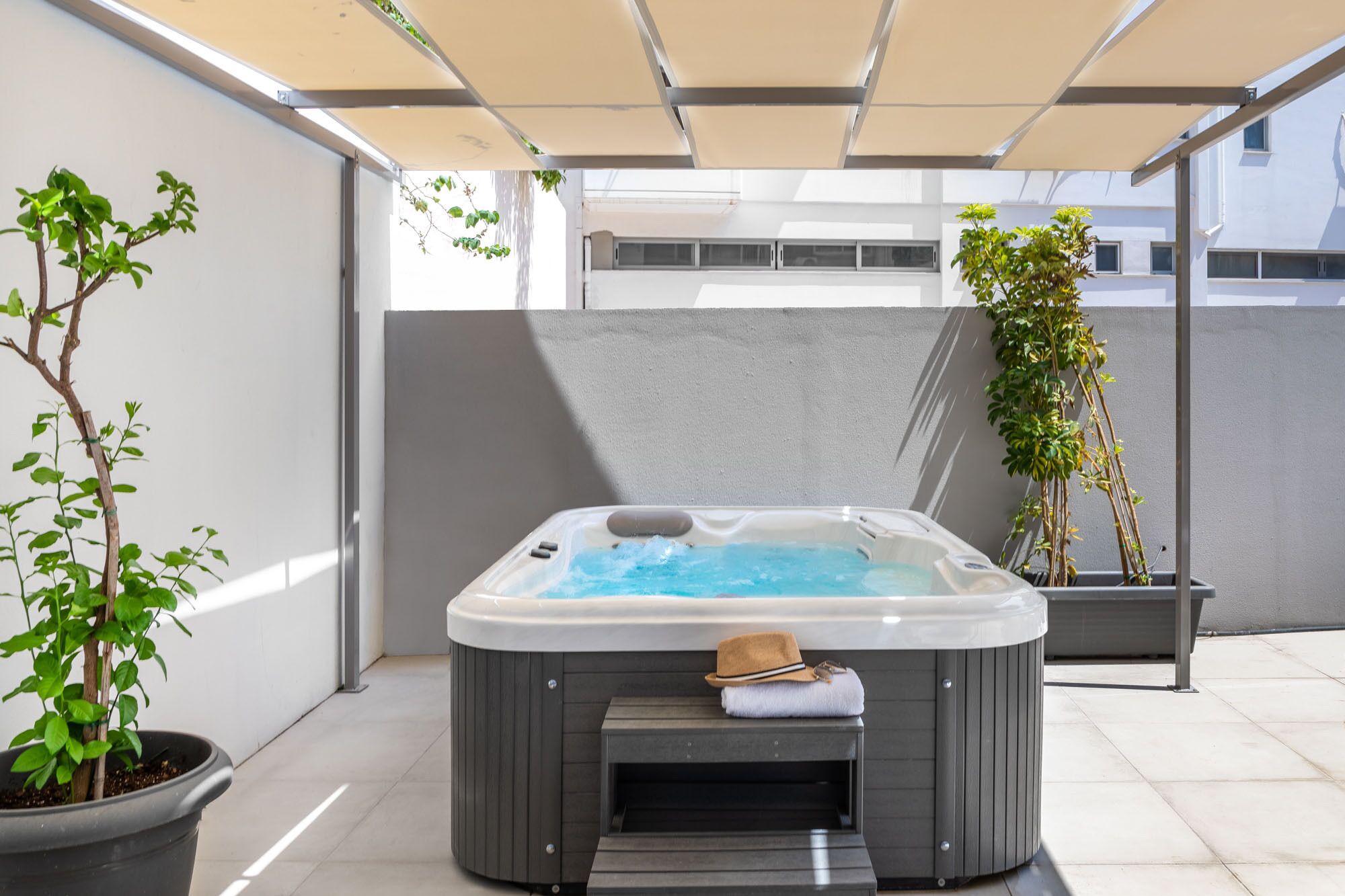 A hot tub in an outdoor setting with a pergola overhead, surrounded by potted plants and a modern building in the background.