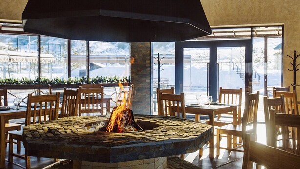A restaurant with a central fire feature, wooden tables and chairs, and large windows offering a view of the outdoors. - Fazenda U Mihalyicha (Talgar)