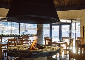Un restaurante con una chimenea central, mesas y sillas de madera, y grandes ventanales que ofrecen vistas al exterior.