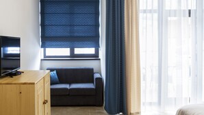 A modern room with a television on a wooden cabinet, a dark sofa, and a large window with blue curtains. - Fazenda U Mihalyicha (Talgar)