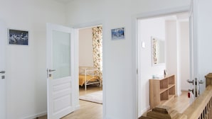 A hallway with a wooden floor, white walls, and a wooden railing. There are doors leading to a bedroom with a bed and a bathroom with a mirror and a wooden cabinet. - Fazenda U Mihalyicha (Talgar)