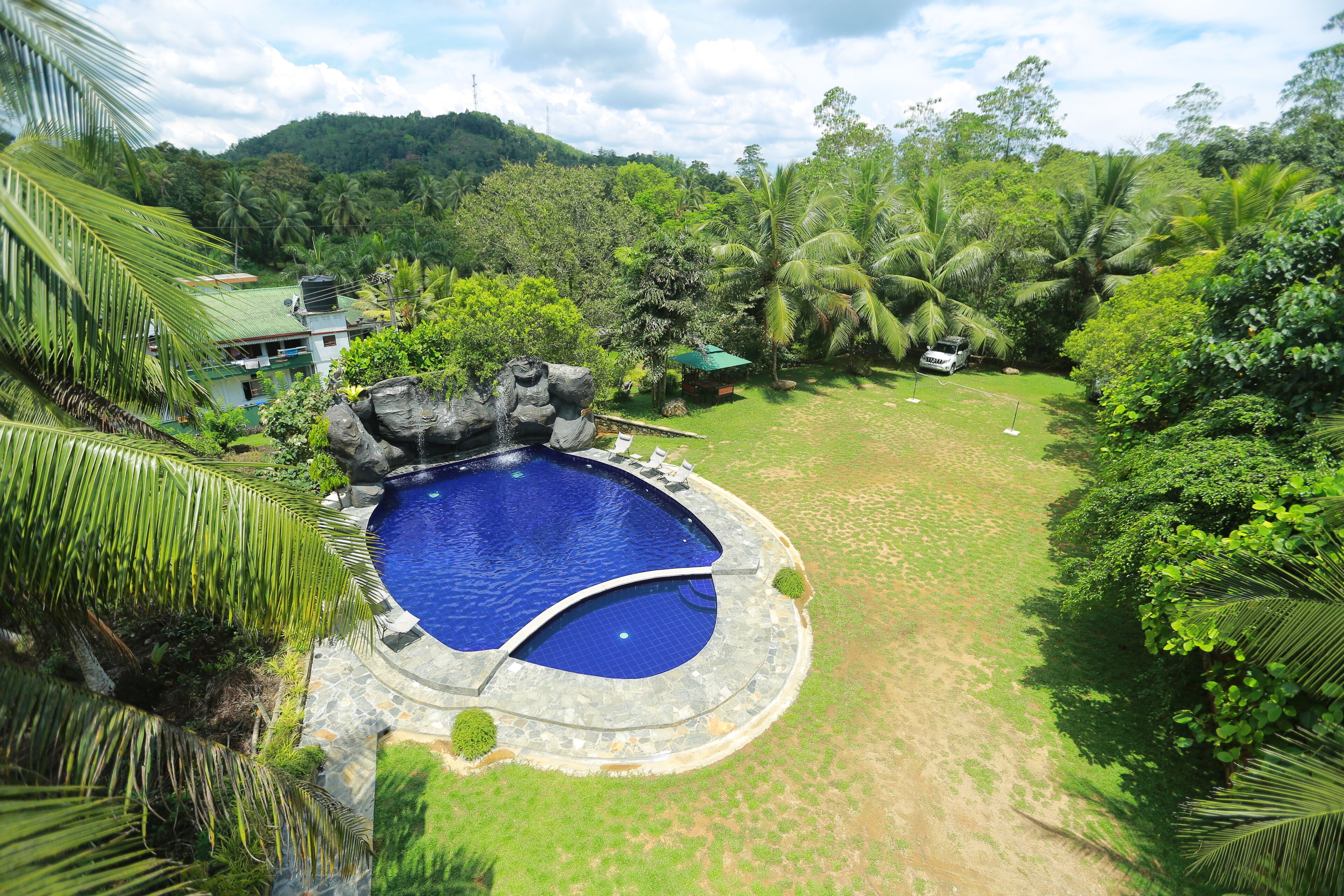 A circular pool with a stone edge, surrounded by lush greenery and palm trees, with a view of a hill in the background.