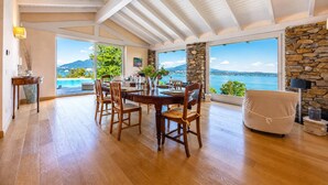 Un vaste espace dédié à la salle à manger, avec une table et des chaises en bois, de grandes fenêtres offrant une vue sur la mer et un mur en pierre.