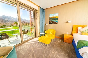 Una habitación de hotel con un ventanal grande, una cama, un sillón amarillo, una mesita pequeña y una mesita de noche.
