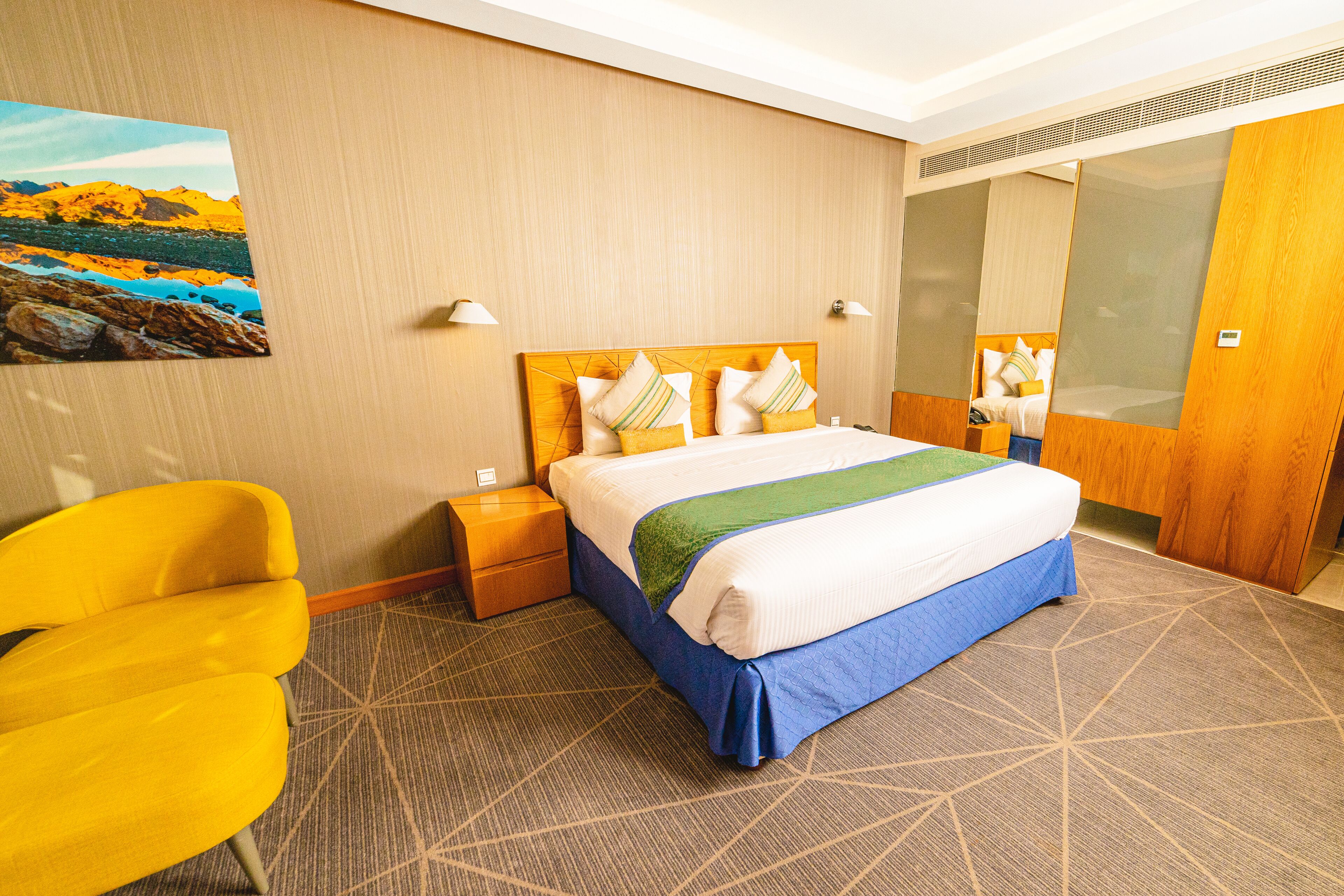 A hotel room with a large bed, a yellow armchair, a nightstand, and a painting on the wall.