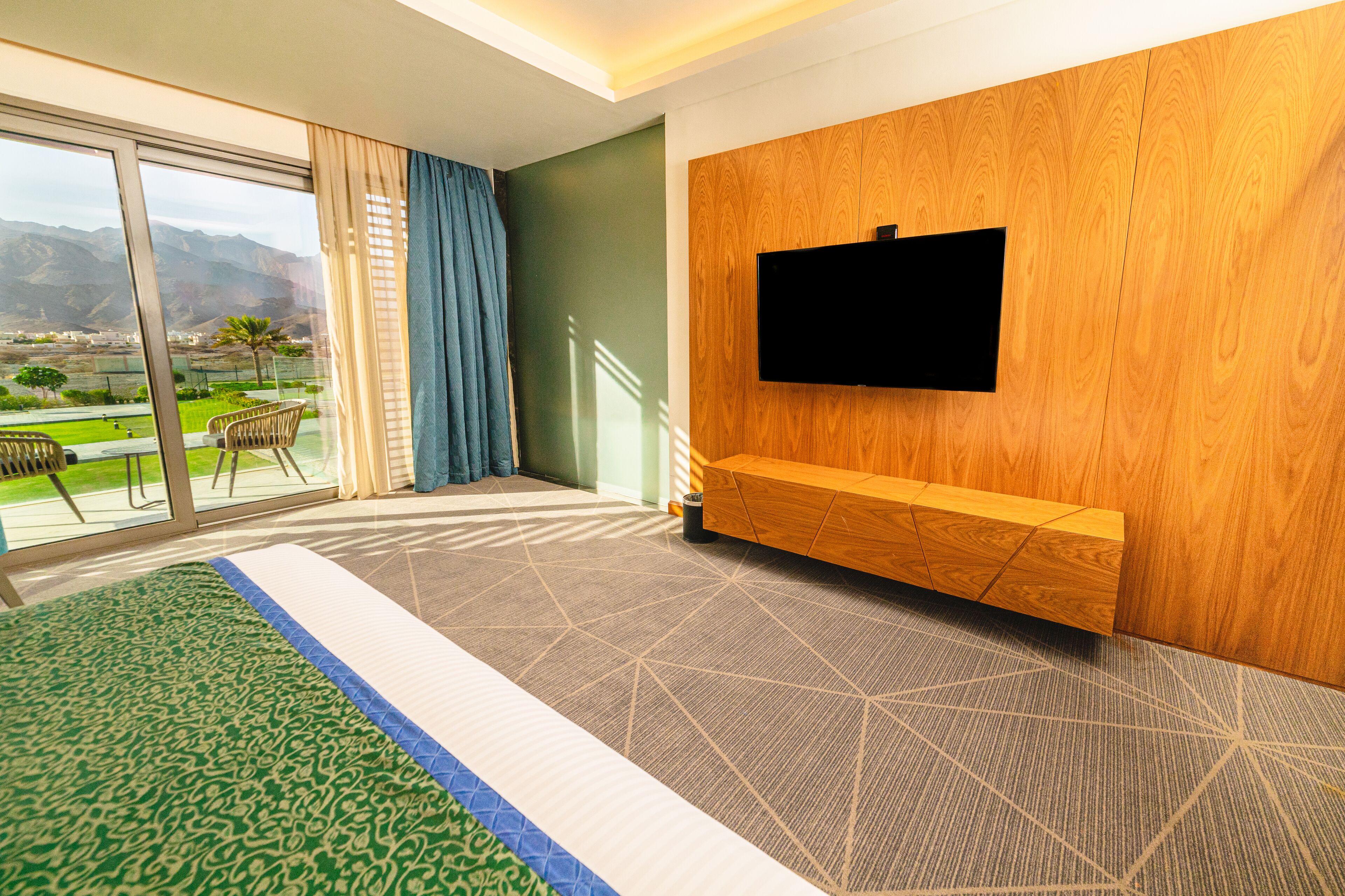Een hotelkamer met een groot raam, een flatscreen televisie bevestigd aan een houten wand, een kamerbreed tapijt en een zithoek met een stoel en tafel.