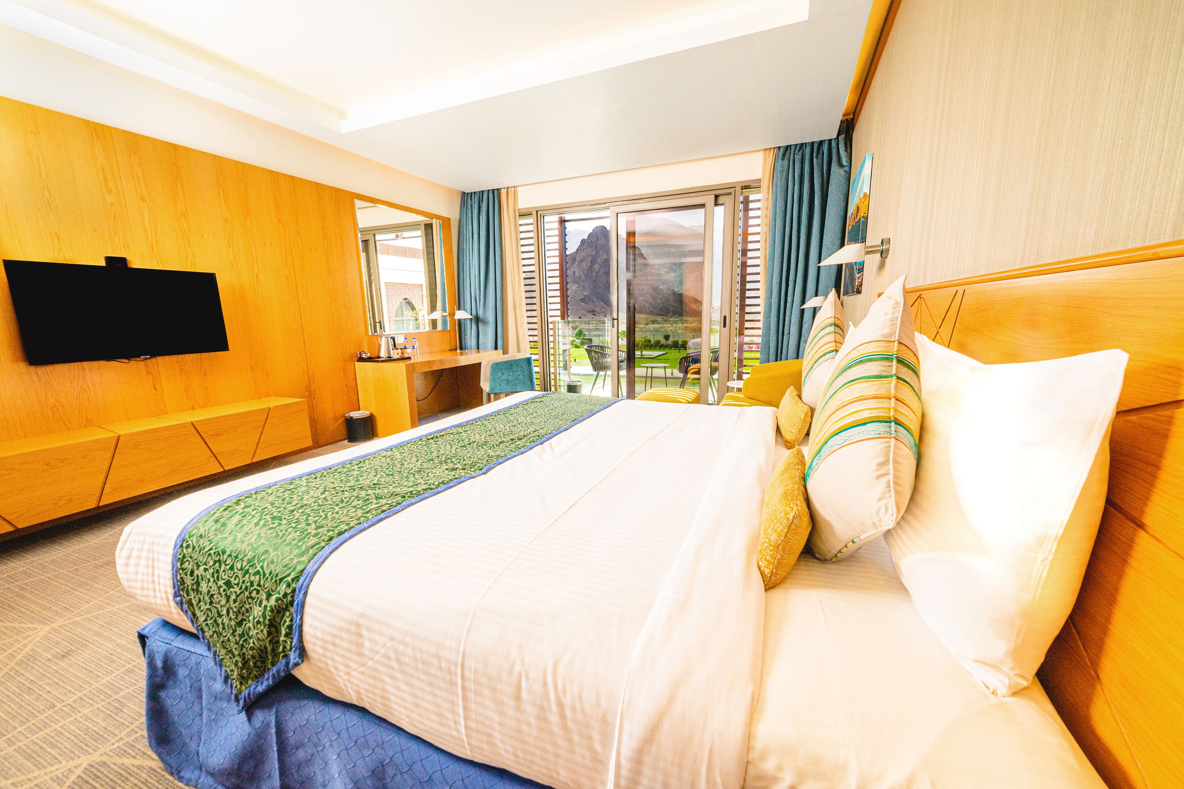 A hotel room with a large bed, a flat-screen TV mounted on the wall, a desk with a chair, and a view of the outdoors through a sliding glass door.
