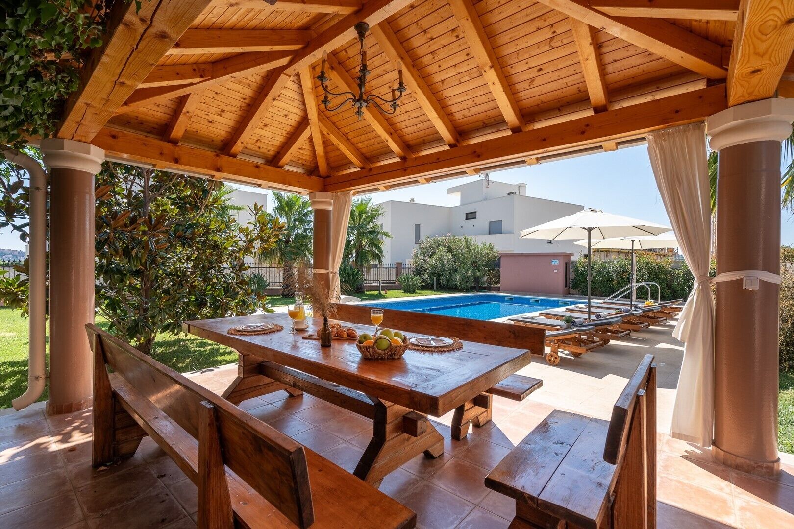 A wooden pergola with a pool table and benches, surrounded by a pool and greenery.