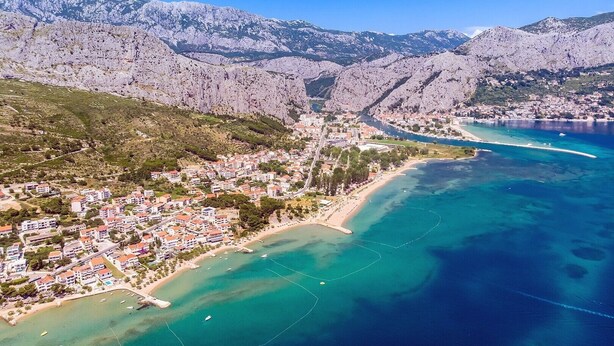An aerial view of a coastal town with a beach, residential buildings, and mountainous terrain in the background.