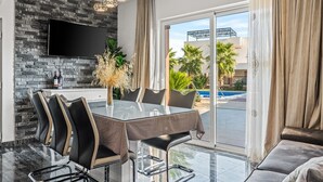 A modern dining area with a glass table, black and beige chairs, a flat-screen TV mounted on a stone wall, and a view of a swimming pool and palm trees through large glass doors.