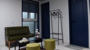 A room with a green sofa, a small table with a vase, two ottomans, a window with blinds, a coat rack, and a blue door. - We Home Guesthouse - Hostel (Incheon)
