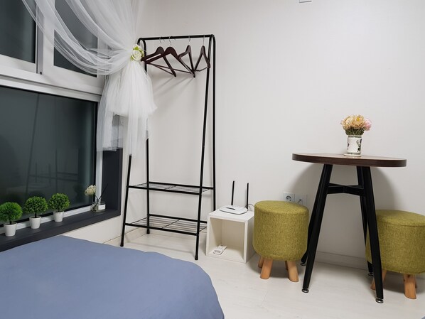 Un dormitorio con una cama, una mesita de noche con un florero, una estantería metálica negra y una ventana con cortinas de encaje.