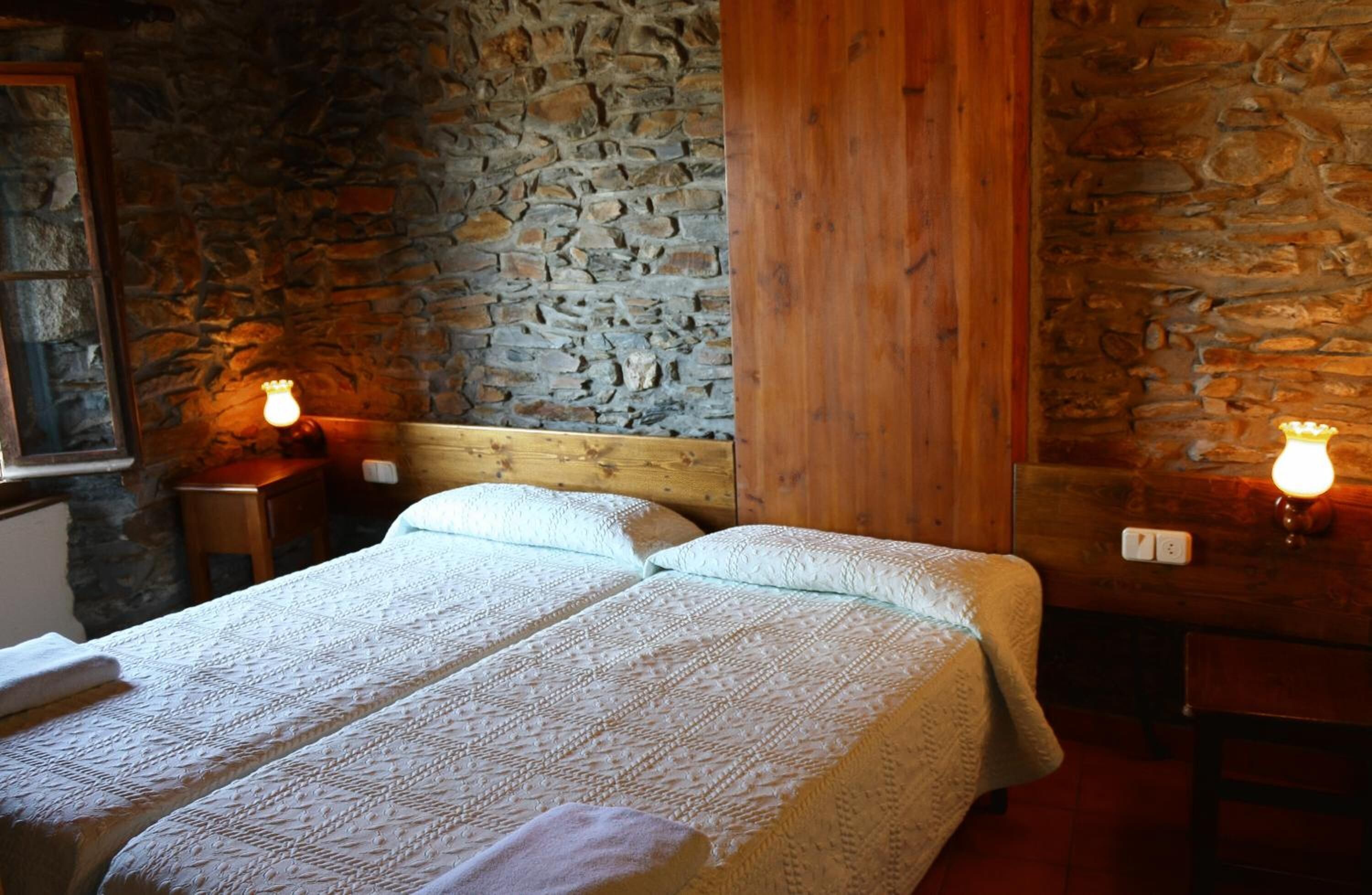 Une chambre avec un lit double doté d’une tête de lit en bois et de deux tables de chevet, chacune avec une lampe. Les murs de la chambre sont en pierre et le lambris est en bois.