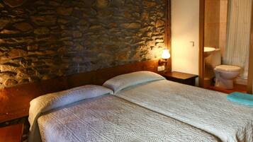 Una habitación con pared de piedra, cama de madera con colcha blanca, mesita de noche con lámpara y baño con inodoro y cortina de ducha.