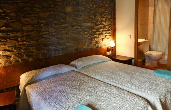 A room with a stone wall, a wooden bed with a white quilt, a nightstand with a lamp, and a bathroom with a toilet and a shower curtain.