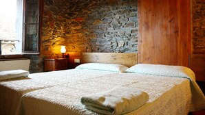 A bedroom with a stone wall, wooden ceiling beams, a bed with a quilt, a nightstand with a lamp, and a window with a view of the outside. - Hostal Ruso (Llívia)
