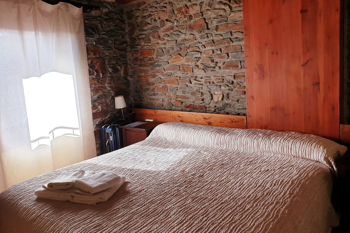 A cozy room with a stone wall, wooden beams, a neatly made bed with a folded towel, and a suitcase on a nightstand.