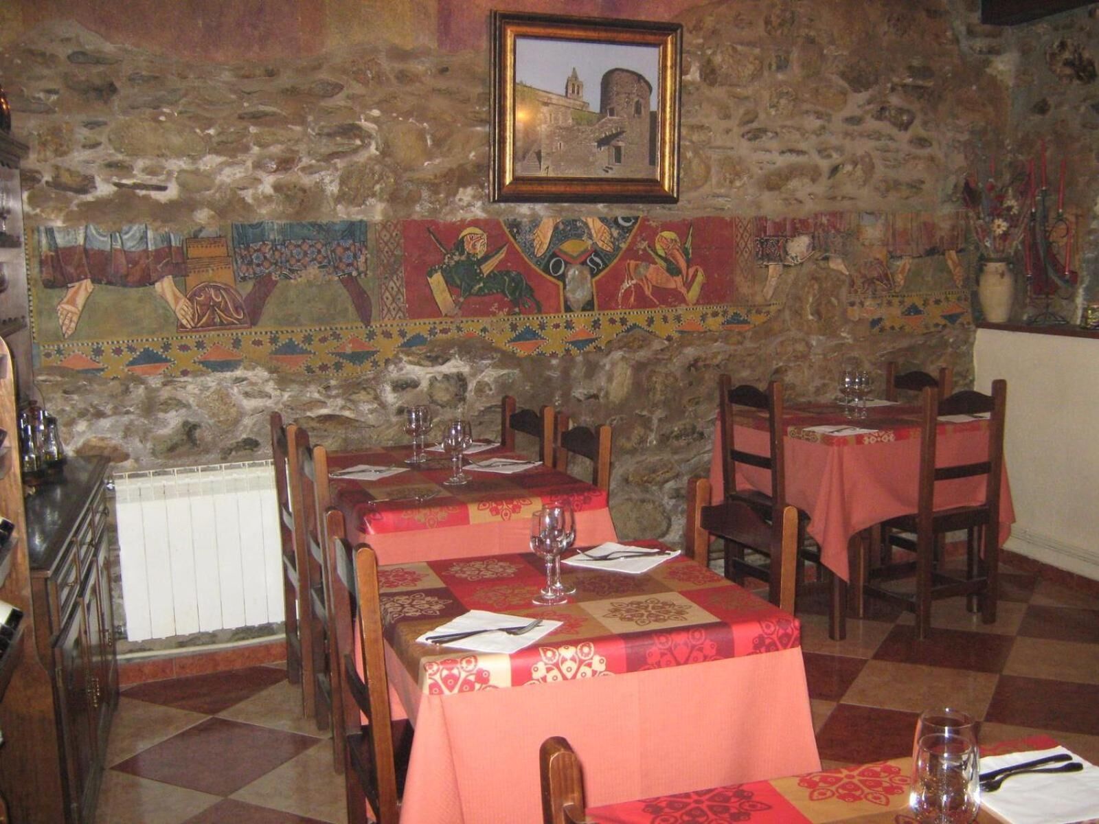 A restaurant with stone walls and a mosaic mural. Tables are set with red tablecloths and wine glasses.