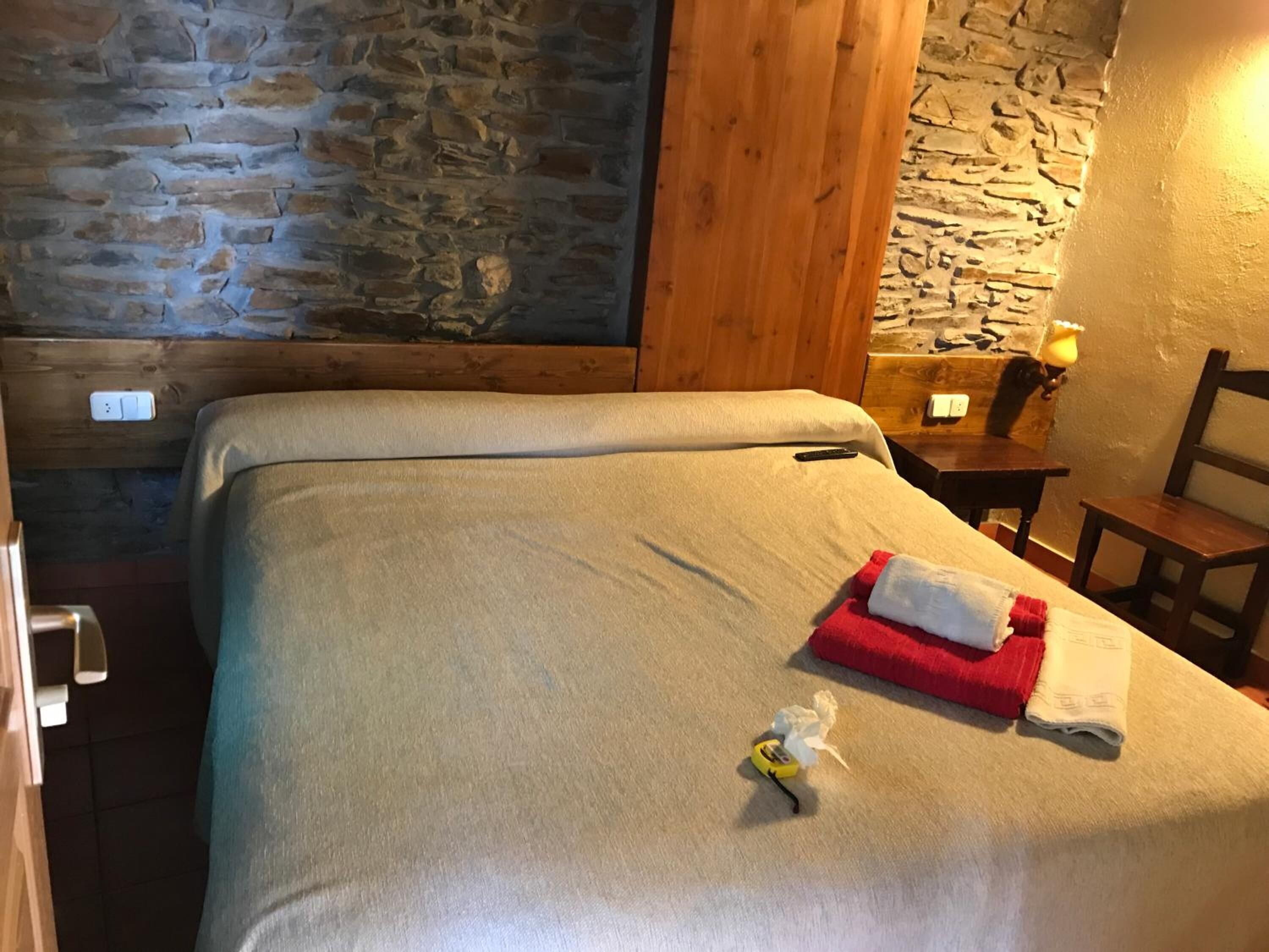 A neatly made bed with a white blanket and a red towel, a small bedside table with a lamp, and a wooden chair.