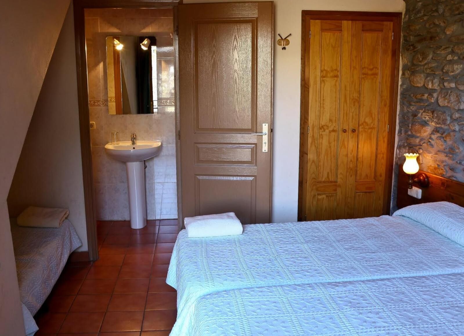 A bedroom with a bed, a wooden door, a bathroom with a sink, and a stone wall.