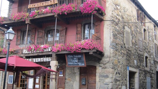 A building with a sign reading "Restaurante de Ventura" and a balcony with pink flowers. There are wooden barrels outside and a red umbrella. - Hostal Ruso (Llívia)