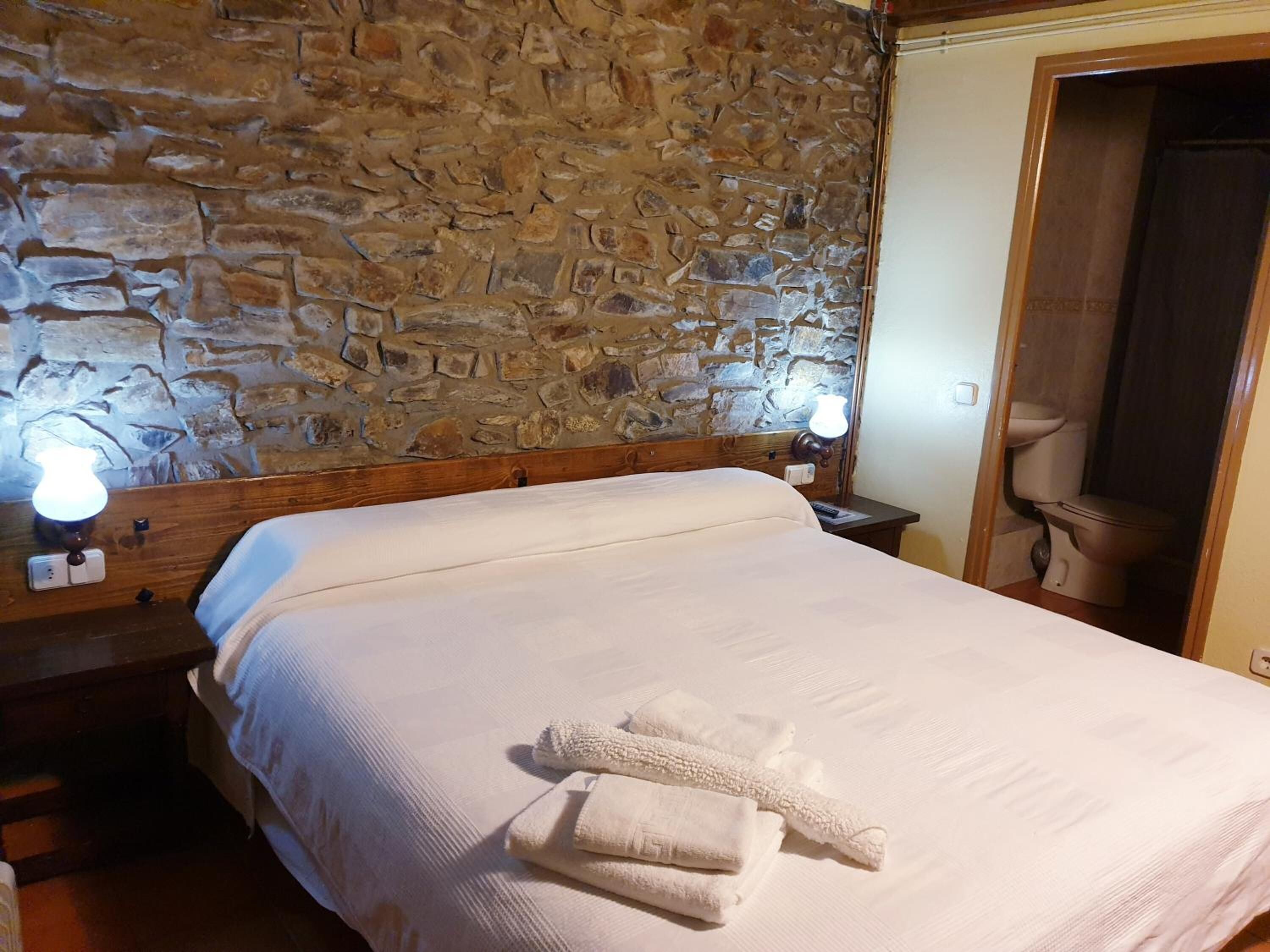 Una habitación con pared de piedra, una cama con ropa de cama blanca, dos lámparas de noche y un baño visible a través de una puerta abierta.