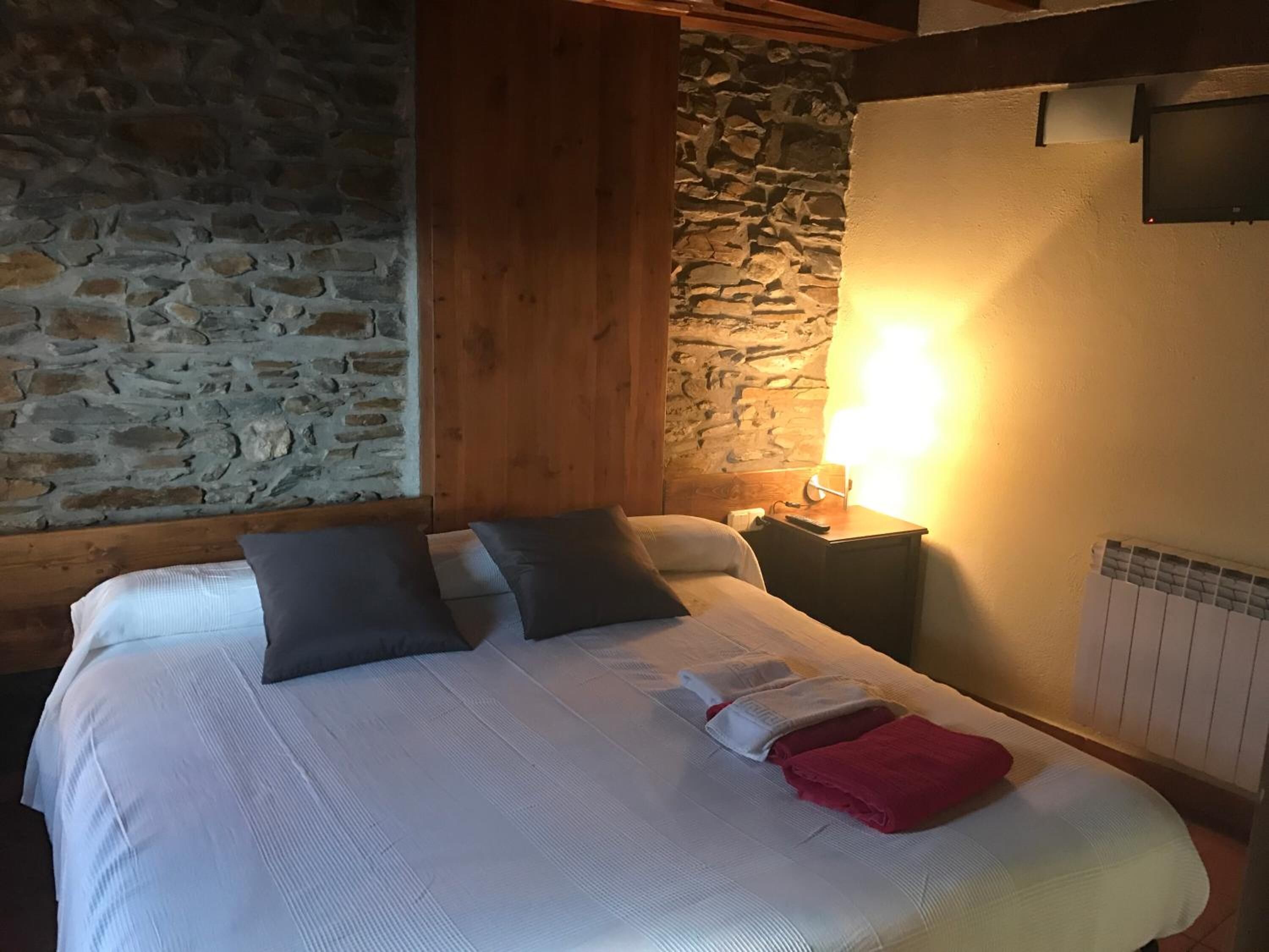 A bedroom with a stone wall, wooden beams, a bed with pillows, a nightstand, and a wall-mounted lamp.