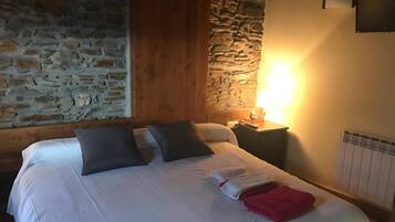 A bedroom with a stone wall, wooden beams, a bed with pillows, a nightstand, and a wall-mounted lamp.
