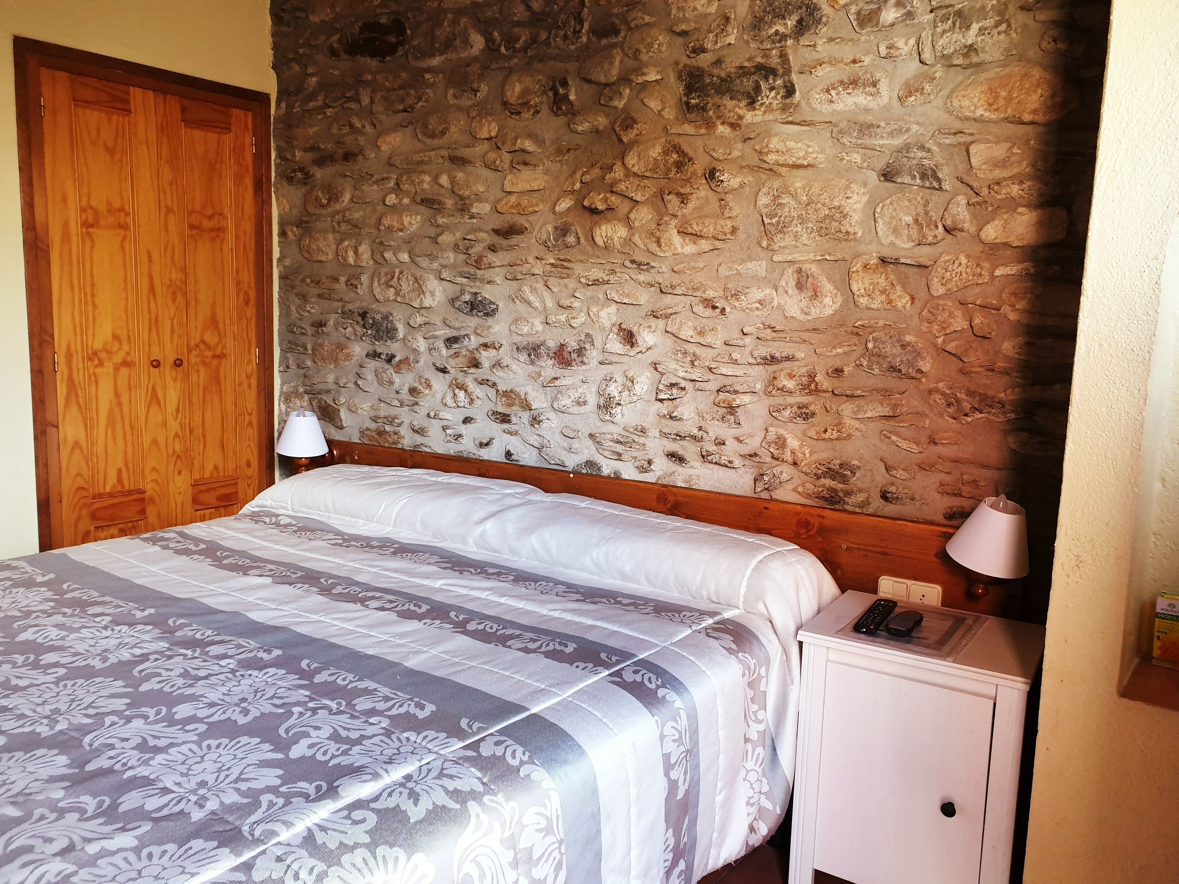 A bedroom with a stone wall, wooden wardrobe, bed with patterned bedding, bedside lamp, and a small cabinet with a phone and remote control.