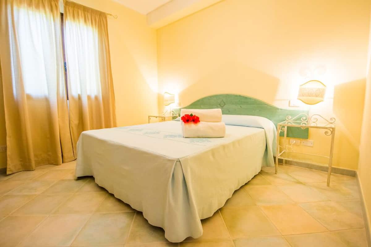 Un lit bien fait, recouvert d’une couvre-lit blanche et agrémenté d’un coussin décoratif, une table de chevet avec une lampe et une fenêtre avec des rideaux.