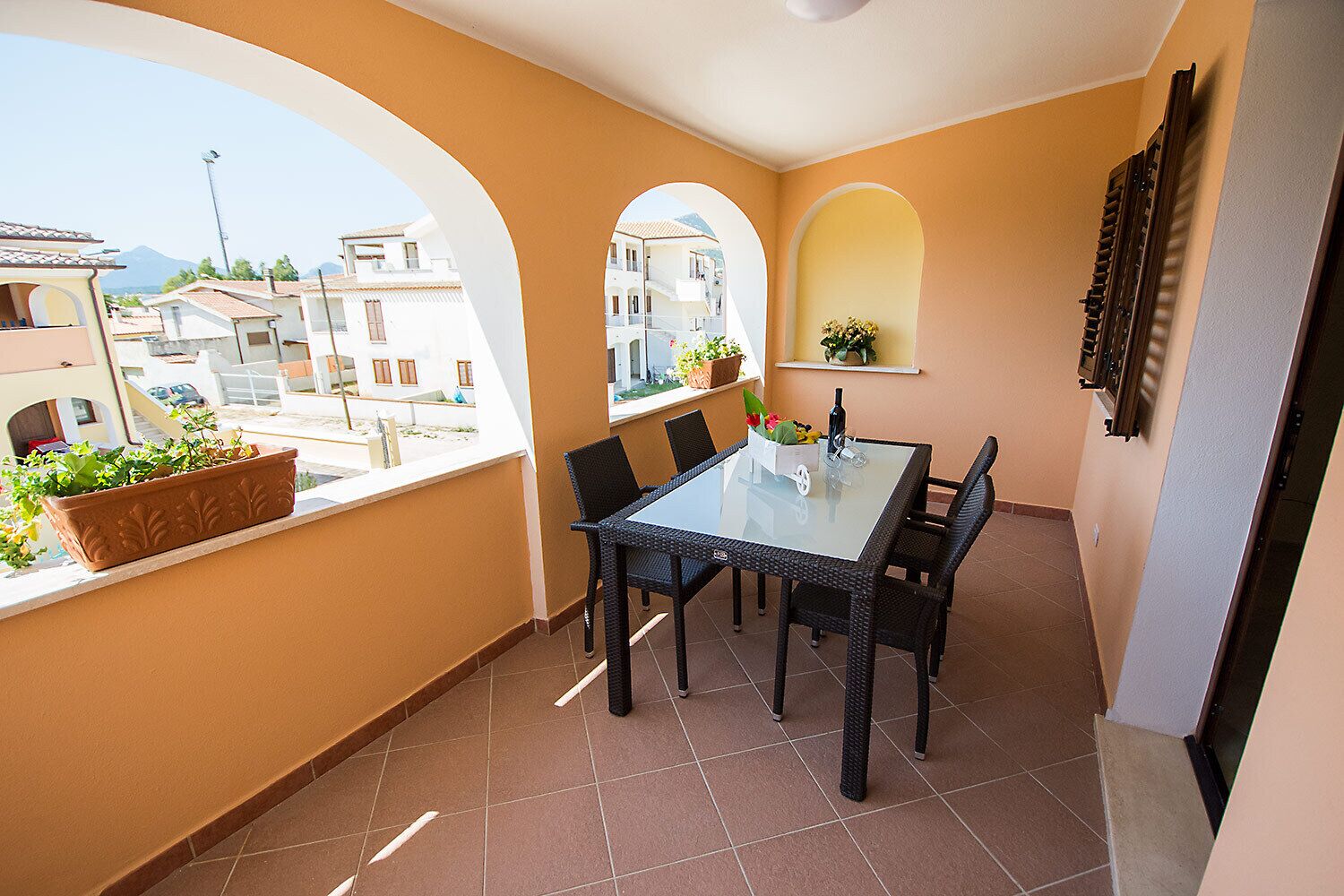 A balcony with a glass table and chairs, potted plants, and a view of buildings.