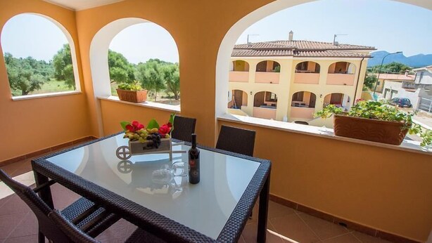 A balcony with a glass table and chairs, potted plants, and a view of a building with arches. - Le Residenze del Maria Rosaria - Vico Friuli 1 (Orosei)