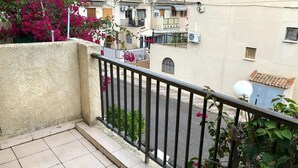 A balcony with a tiled floor, a metal railing, and a view of a street with buildings and a clear sky.